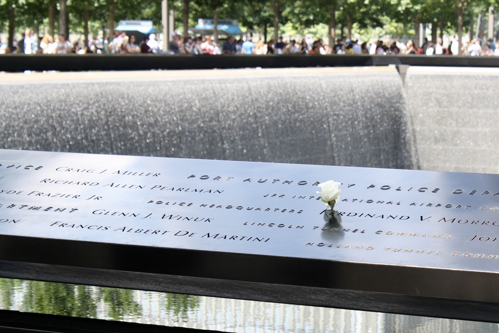 A white rose is placed on the 9/11 memorial, through the name of one of the victims. In the background, a fountain pours water into a deep opening.